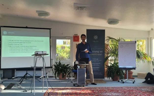 Referent Dr. Chaban Salih hält seinen Vortrag zu Kommunikation und PR vor einer Präsentation, daneben das Roll-up der Amana Akademie.