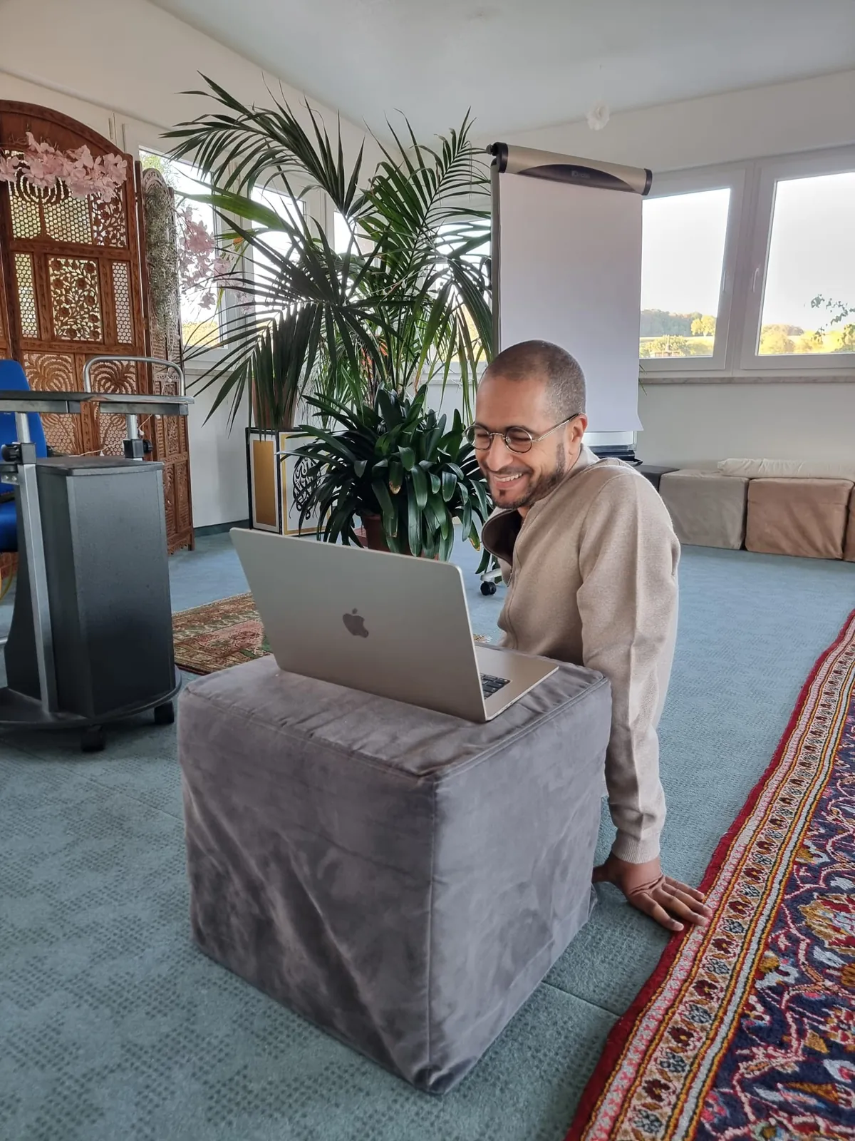 Nadir lacht bei der Vorbereitung am Laptop, der auf einem Sitzwürfel im Seminarraum steht.