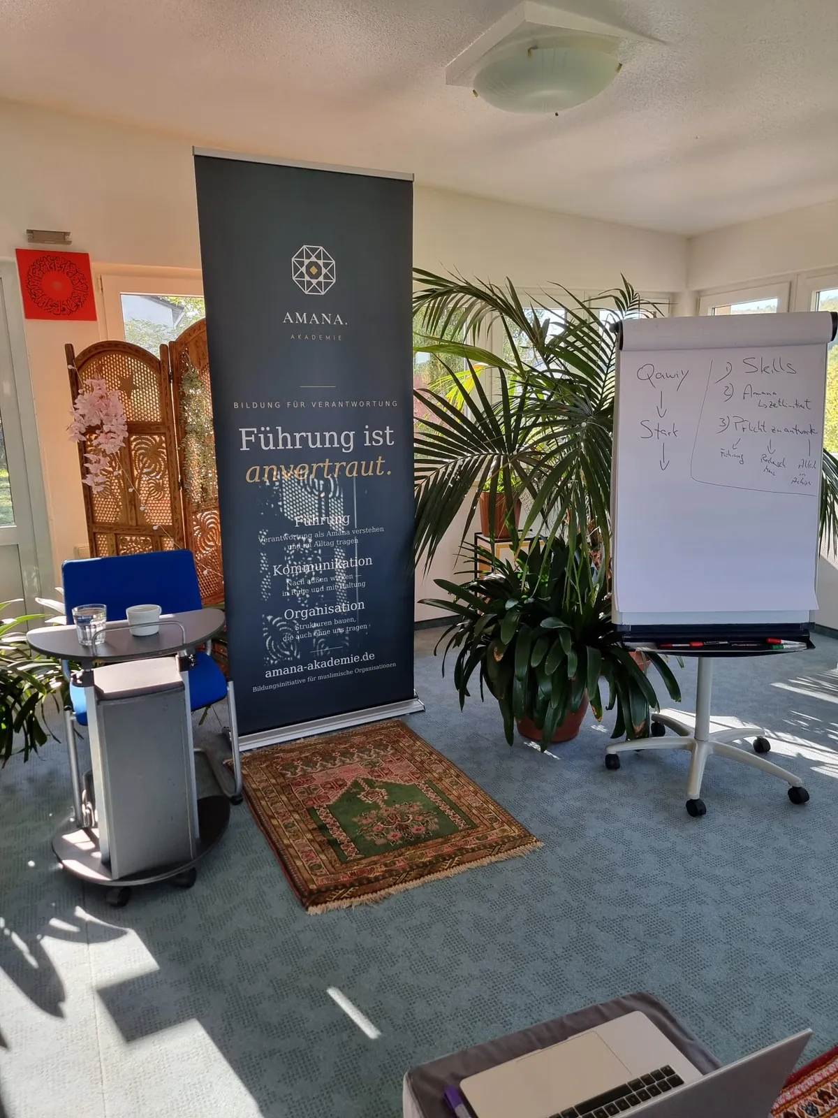 Roll-up der Amana Akademie mit dem Leitsatz »Führung ist anvertraut.«, daneben ein Flipchart mit Stichworten zur Leadership-Einheit und ein Gebetsteppich.